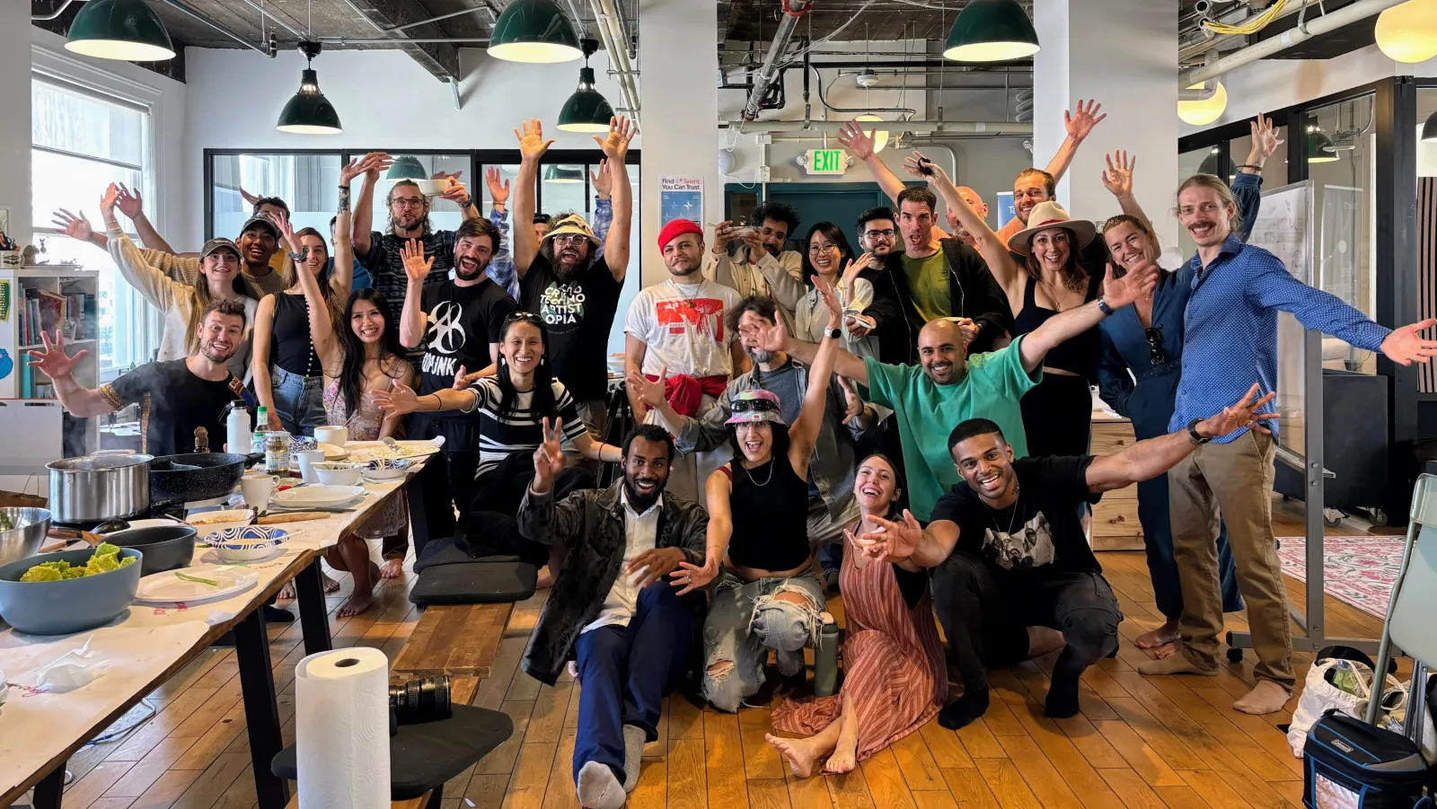 About thirty people cheering with arms raised around long tables set with pots and bowls in an open loft space